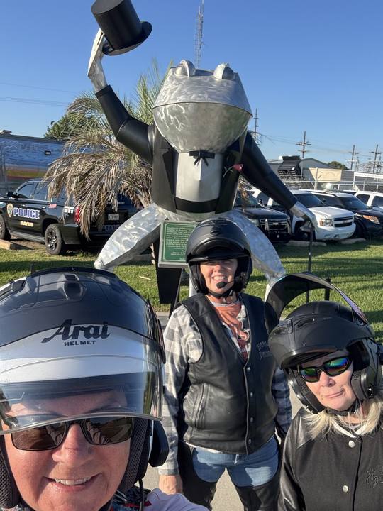 Three riders in helmets taking a photo in front of a tall frog statue